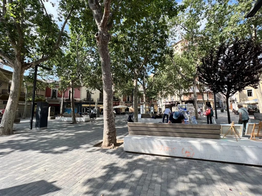 Plaza arbolada en el barrio de Horta, Barcelona, con bancos y comercios.